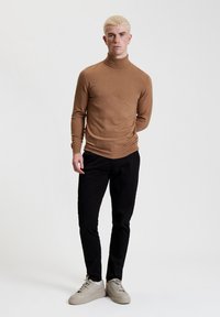 Maglione a collo alto marrone, design aderente, abbinato a pantaloni neri slim-fit e sneaker color crema. Texture liscia, accenti minimali.