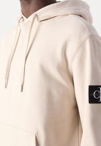 Béžová mikina s kapucí na stahování, klokankou a černou nášivkou na rukávu. Měkká látka a minimalistický design.