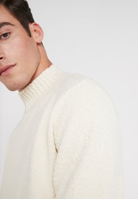 Suéter blanco roto hecho de un tejido de punto texturizado, con un alto cuello acanalado y un corte relajado. La vista lateral enfatiza el material suave.