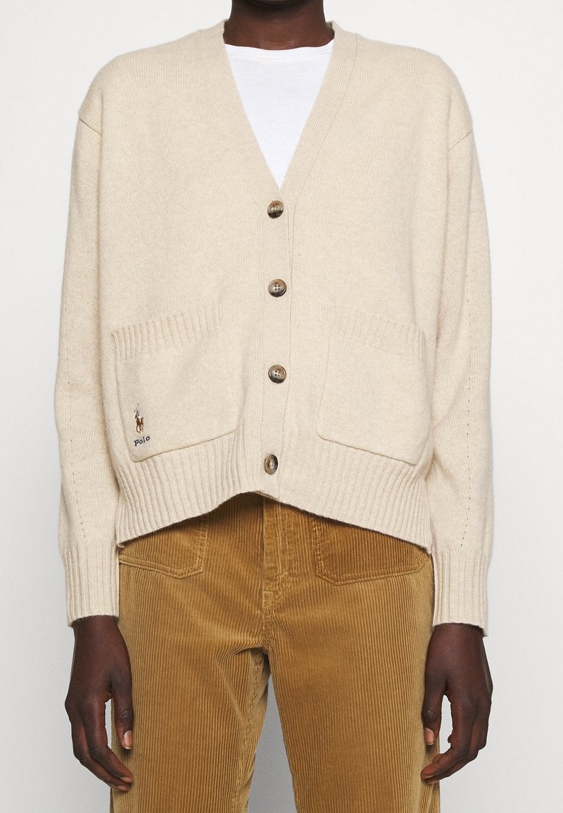 Beige pletený cardigan s výstřihem do V, vybavený žebrovanými manžetami, dvěma předními kapsami a hnědými knoflíky. Akcent loga Polo na levé kapse.