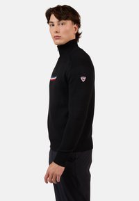 Schwarzer Strickpullover mit hohem Kragen, der einen horizontalen roten, weißen und blauen Streifen über die Brust und ein Logo-Patch am Ärmel aufweist.