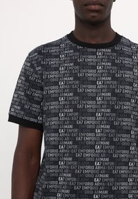Černé a šedé tričko s krátkým rukávem, na kterém je opakující se text "EA7 Emporio Armani", kulatý výstřih a minimální textura.