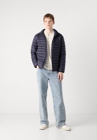Navy puffer jacket with quilted texture, zip closure, and logo patch, worn over a beige polo and paired with light blue baggy jeans.