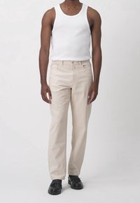 Pantaloni di cotone beige con taglio dritto, dotati di tasca frontale, cuciture a vista, indossati con una canottiera bianca e scarpe nere.