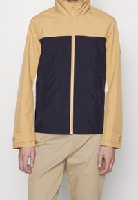 Man som bär en beige och svart zip-up jacka med beiga byxor, stående mot en vit bakgrund.