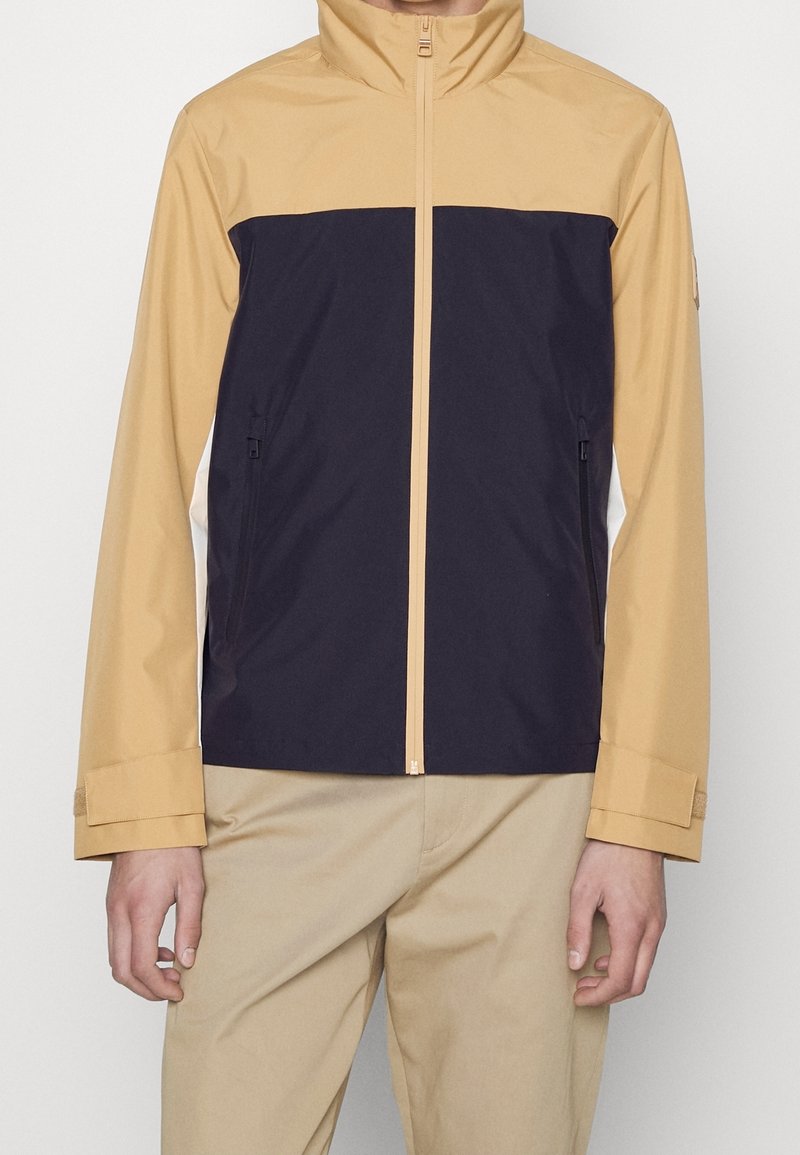 Man som bär en beige och svart zip-up jacka med beiga byxor, stående mot en vit bakgrund.