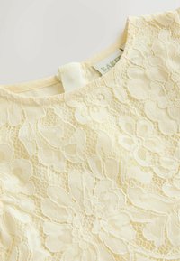 Kollane pitsiga topp, millel on lillemustrid, keerukad detailid ja sile tekstuur. Silt on nähtav kaelas.