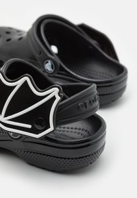 Des Crocs noirs avec une surface texturée, des perforations pour la respirabilité, et un élément décoratif en forme de feuille blanche contrastante sur la sangle.