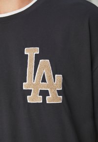 Camiseta gris oscuro que presenta un gran logo bordado en color beige de "LA" con un acabado texturizado y suave. Ribete blanco alrededor del escote.