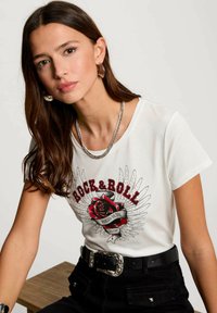 T-shirt di cotone bianco con una graphic di rosa rossa con ali e il testo "ROCK & ROLL". Indossata con pantaloni neri e una cintura con fibbia argentata.