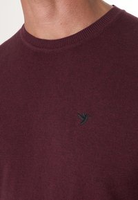 Maroon knitted sweater with a round neckline and a small black bird logo on the left chest. Smooth texture and ribbed collar details.