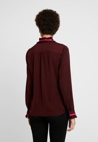 Camicia bordeaux a maniche lunghe con colletto arricciato e accenti rosa a contrasto sui polsini, caratterizzata da una vestibilità rilassata e un dettaglio arricciato nella parte posteriore.