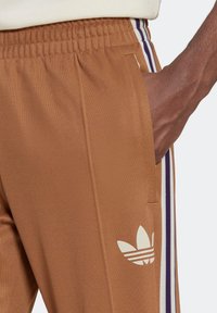 Pantalones deportivos marrones de tela suave con cintura elástica, que presentan el logo blanco de Adidas y rayas laterales moradas como detalle.
