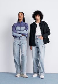 To modeller står side om side. Den ene har på seg en lilla og blå zip-up jakke og lyse denim jeans. Den andre har på seg en svart skjortejakke og lyse jeans.