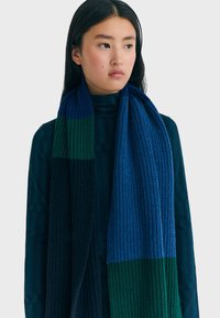 Écharpe en tricot avec des rayures bleues, vertes et bleu marine foncé. Design en côtes texturées, longue et enroulée autour du cou.