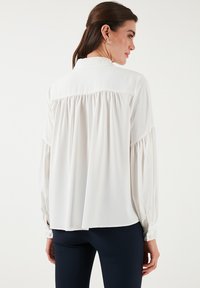 Bílá, lehká blůza s uvolněným střihem, detail akorát na zádech a dlouhými rukávy s knoflíkovými manžetami. Hladká tkanina, minimalistický design.