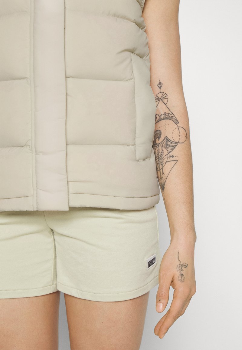 Beige Steppweste mit gestepptem Design, ausgestattet mit einem Reißverschluss vorne und seitlichen Taschen, getragen über hellgrünen Shorts. Tattoo am Arm sichtbar.