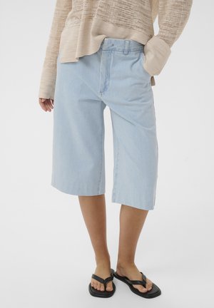 Személy világoskék térdig érő farmer rövidnadrágot, bézs texturált hosszú ujjú felsőt és fekete, négyzet alakú orrú szandált visel, egyik kezével a zsebében áll.