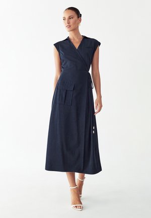 Vrouw draagt een mouwloze marineblauwe wikkeljurk met een borstzak, een grote zijzak en witte enkellaarsjes met enkelbandje, staand op een witte achtergrond.