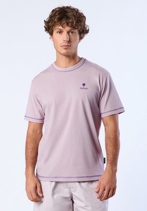 Jeune homme aux cheveux bouclés portant un t-shirt FAGUO violet clair et un pantalon assorti de couleur claire, debout devant un fond uni.