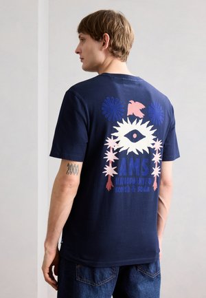 Muž nosí tmavomodré tričko s veľkým grafickým motívom oka, kvetín a textu „AMS handpainted scotch & soda“ na chrbte.