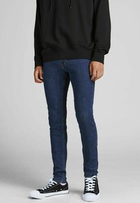 Man wearing dark blue skinny jeans, black hoodie, and black low-top sneakers with white soles and laces, standing on gray background.