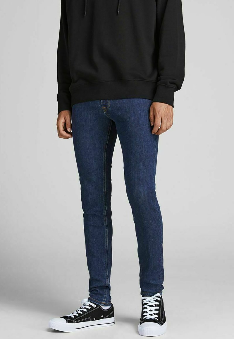 Man wearing dark blue skinny jeans, black hoodie, and black low-top sneakers with white soles and laces, standing on gray background.