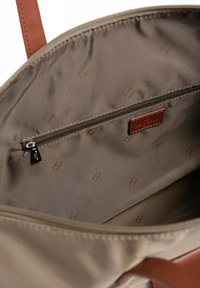 Interno della borsa in canvas con una fodera liscia marrone, un taschino con zip e un'etichetta in pelle. I loghi decorativi sono stampati in modo discreto sul tessuto.