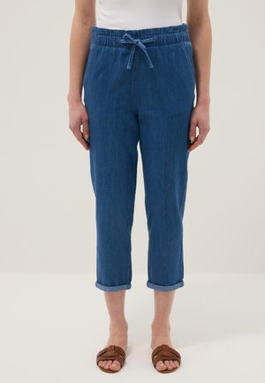 Woman wearing rolled-up blue denim jogger pants with a drawstring waist and brown braided open-toe sandals.