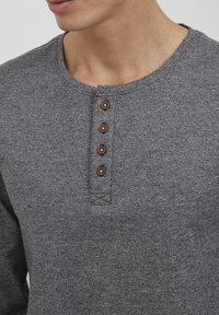 Grå langermet henley-skjorte med rund hals og fem brune knapper. Stoffet har en teksturert, lett melert utseende.