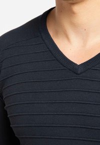 Detailní záběr na osobu v tmavě modrém svetru s výstřihem do V, s horizontálně žebrovanou texturou přes hrudník a ramena.