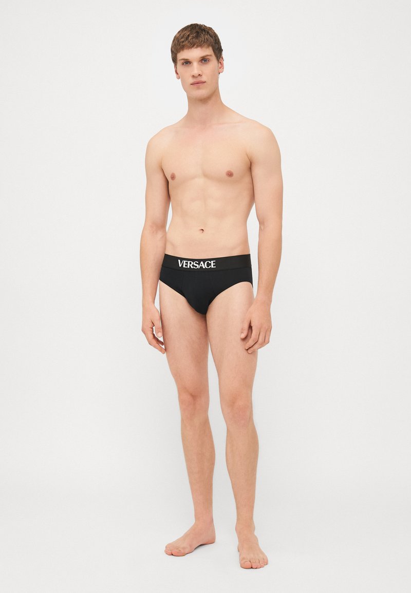 Boxers noirs Versace avec un logo blanc sur la ceinture, design ajusté et texture lisse. Modèle stand pieds nus, fond neutre.