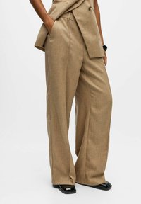 Pantalons beige à jambes larges en tissu texturé, avec une taille ceinturée et des poches inclinées, associés à des chaussures noires brillantes.