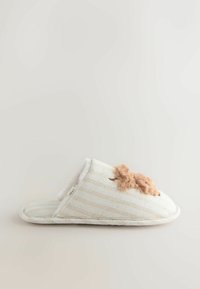 Zapatilla de rayas color crema hecha de tela con un detalle de piel de tono marrón claro y suave. Cuenta con un interior acolchado y una suela plana.