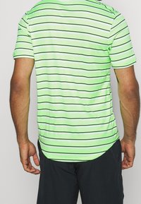 Hombre vistiendo una camiseta polo de color verde claro con finas rayas horizontales en negro y blanco, combinado con pantalones cortos negros, visto desde atrás.
