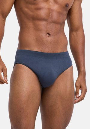 Spierige man in donkerblauwe slip, staande tegen een effen witte achtergrond, torso en bovenbenen zichtbaar.