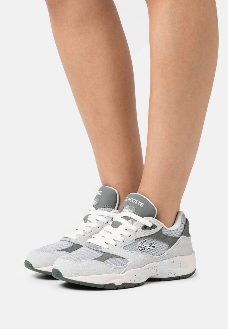 Zapatillas grises y blancas con materiales de malla y ante, con acentos verdes, suela estriada y un logotipo prominente en el lateral.