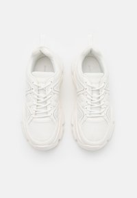 Bílé sportovní boty mají svrchní část z prodyšné síťoviny, zaoblenou špičku a silnou gumovou podešev. Šňůrky a poutka zvyšují jejich funkčnost.