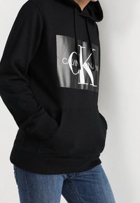 Felpa nera con cappuccio, caratterizzata da un grande logo bianco di Calvin Klein in un design rettangolare. Il materiale sembra morbido e presenta una tasca frontale.