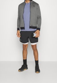 Chaqueta deportiva gris con cremallera, camiseta morada, pantalones cortos negros con forros blancos, calcetines negros y zapatillas negras con acentos en marrón, sosteniendo una botella de agua.