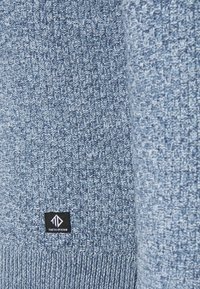 Gros plan sur un tissu tricoté bleu texturé avec une petite étiquette noire portant l'inscription "Tom Tailor Denim" cousue près du bord inférieur.