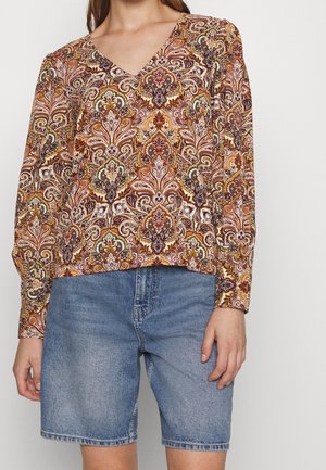 Kvinde iført langærmet V-halsbluse med indviklet orange og brun paisleymønster kombineret med midtlange blå denimshorts.