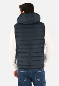 Gilet puffer blu navy con cappuccio, texture trapuntata e aperture elastiche per le braccia, indossato sopra una maglietta bianca a maniche lunghe, abbinato a jeans blu.