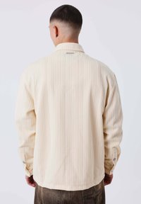 Chaqueta color crema con un patrón de canalé texturizado, mangas largas, cuello y puños con botones, que presenta una etiqueta de marca en la parte posterior del escote.