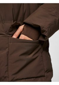 Brūns izolēts žakets ar mīkstu tekstūru, ar roku sānu kabatā, ribotām piedurknēm un gludu apdari.