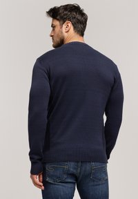 Granatowy sweter dzianinowy z prążkowanym kołnierzem i mankietami, o teksturowanym wykończeniu i prostym dolnym końcu. Nosi się go na dżinsach.