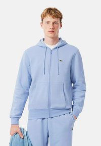 Hoodie zippé bleu clair en tissu doux avec une poche avant, capuche à cordon réglable et logo vert Lacoste sur la poitrine.