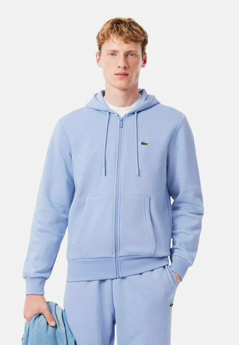 Hoodie zippé bleu clair en tissu doux avec une poche avant, capuche à cordon réglable et logo vert Lacoste sur la poitrine.