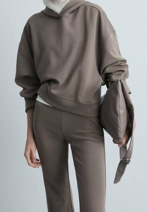 Personne portant un sweat à capuche taupe et un pantalon assorti, tenant une pochette matelassée taupe avec une bandoulière sur un fond uni.