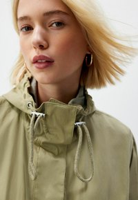 Jachetă de culoare khaki cu guler înalt, șnururi ajustabile și accesorii din metal argintiu. Materialul pare ușor și neted.